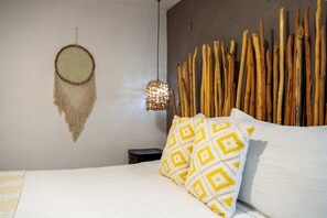 En seng med puder i hvidt og gult mønster, en vævet drømmefanger og en vægmonteret lampe.