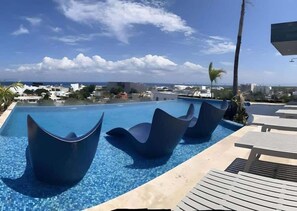 A pool with modern, sculptural chairs and a view of the ocean and buildings. - Ground-Floor Studio · Terrace & Rooftop Pool (Playa del Carmen)