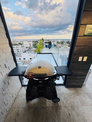 Een kamer met uitzicht op een stadsbeeld door een raam, een scooter tegen de muur geparkeerd, en een houten paneel met een bordje.