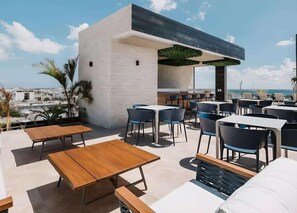 An outdoor seating area with tables and chairs, a modern building structure, and a view of the cityscape. - Ground-Floor Studio · Terrace · Near 5th Ave (Playa del Carmen)