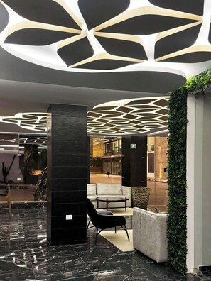 Eine moderne Lobby mit einem geometrischen Deckendesign, einer Sitzecke mit Sofa und Sesseln sowie einer mit Grünpflanzen bepflanzten Wand.