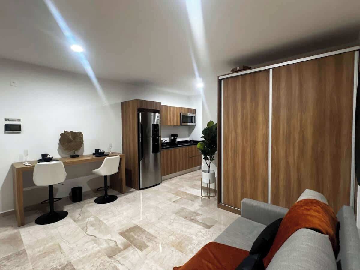 Una cocina moderna con refrigerador, microondas y armarios de madera. Hay un comedor con dos sillas y una mesa, y un sofá en la sala.