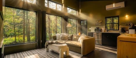 Un interior moderno con sofá, reposapiés y zona de cocina, ventanas amplias con vistas a un bosque e iluminación colgante.