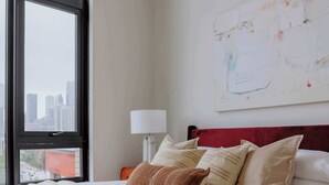Quarto de hotel com uma grande janela, cama com várias almofadas, mesa de cabeceira com abajur e parede com uma obra de arte abstrata.