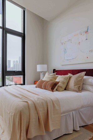 Ein Hotelzimmer mit großem Fenster, einem Bett mit vielen Kissen, einem Nachttisch mit Lampe und einer Wand mit einem abstrakten Kunstwerk.