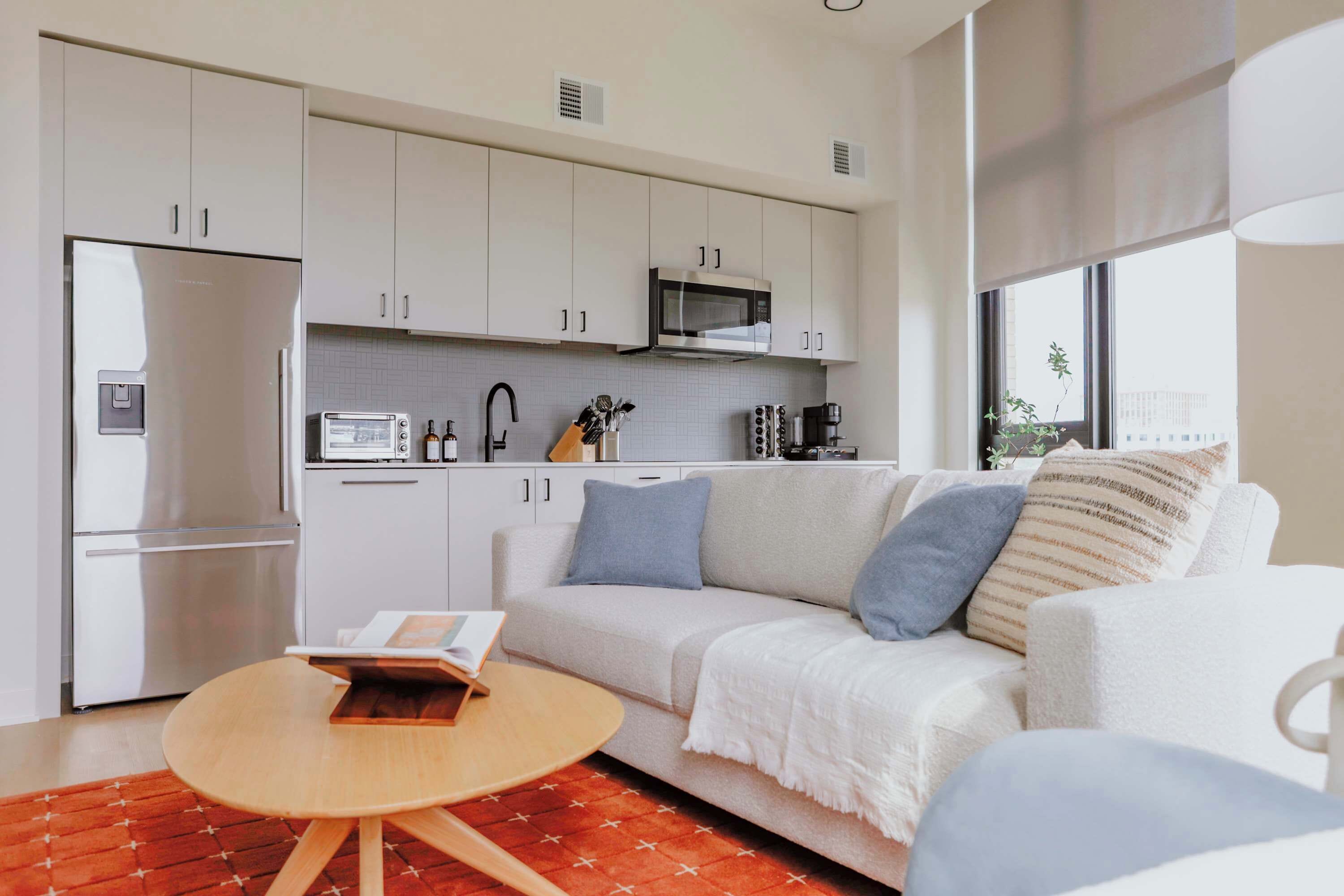 Una cocina moderna con armarios blancos, un microondas y un refrigerador. Hay un sofá blanco con cojines azules y una pequeña mesa de madera con un libro.