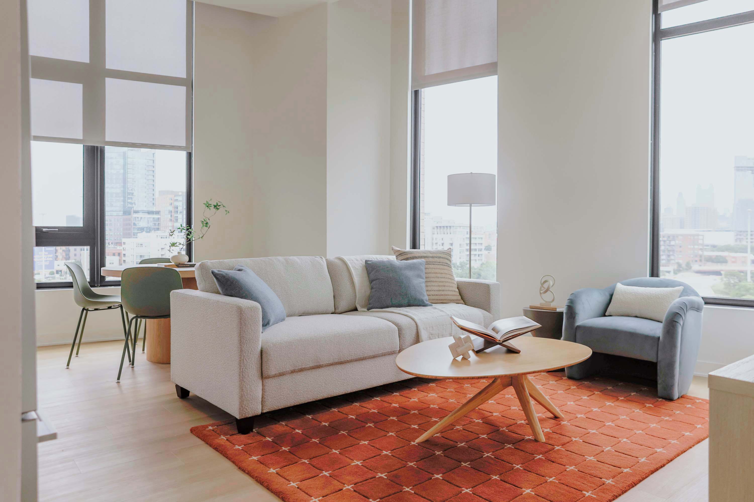 Una sala moderna con un sofá, un sillón, una mesita de centro y vistas de la ciudad a través de grandes ventanales.