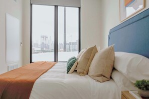 A hotel room with a bed, a view of the city through a large window, and a blue headboard. - Luxury Penthouse Collection PH2 - Fulton Market (Chicago)