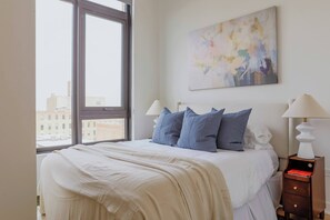 A bedroom with a large window, a bed with white bedding, two bedside lamps, and a painting on the wall. - Luxury Penthouse Collection PH3 - Fulton Market (Chicago)