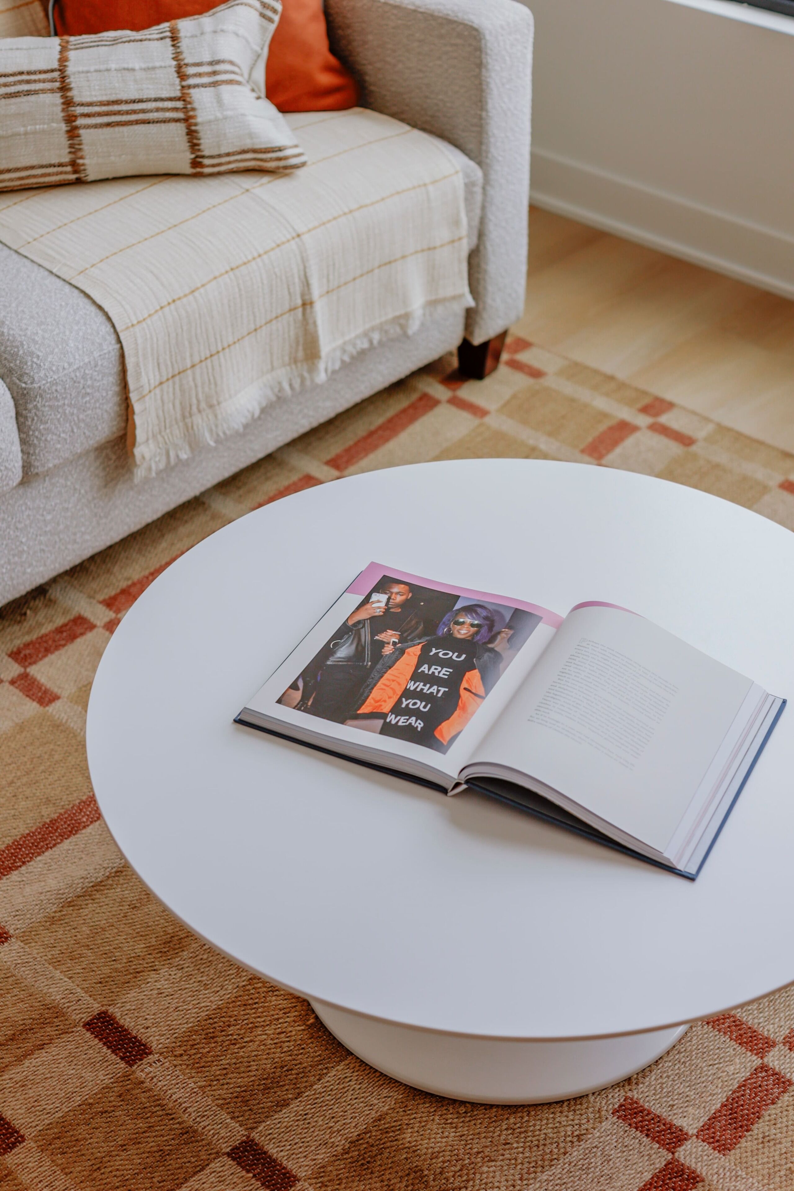 A magazine on a white round table in a room with a patterned carpet and a grey sofa with a throw blanket.