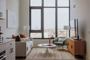 A modern living room with a sofa, armchair, coffee table, TV, and a view of the city through large windows. - Luxury Penthouse Collection PH3 - Fulton Market (Chicago)