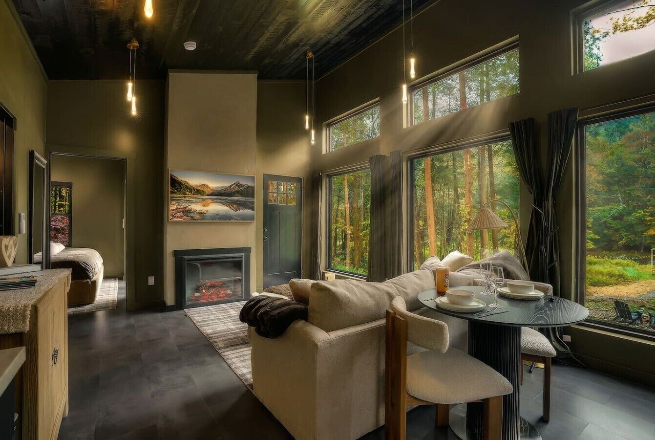 A modern living room with a fireplace, a sofa, a dining table, and large windows with a forest view.