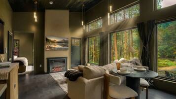 A modern living room with a fireplace, a sofa, a dining table, and large windows with a forest view.