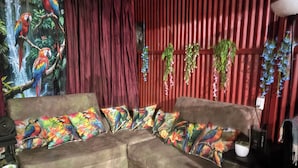 A room with a brown sofa, colorful pillows, a mural of parrots, a red curtain, and hanging plants. - Cottage with a pool and natural surroundings—just 20 minutes from the beaches and Manuel Antonio Park! (Naranjito)