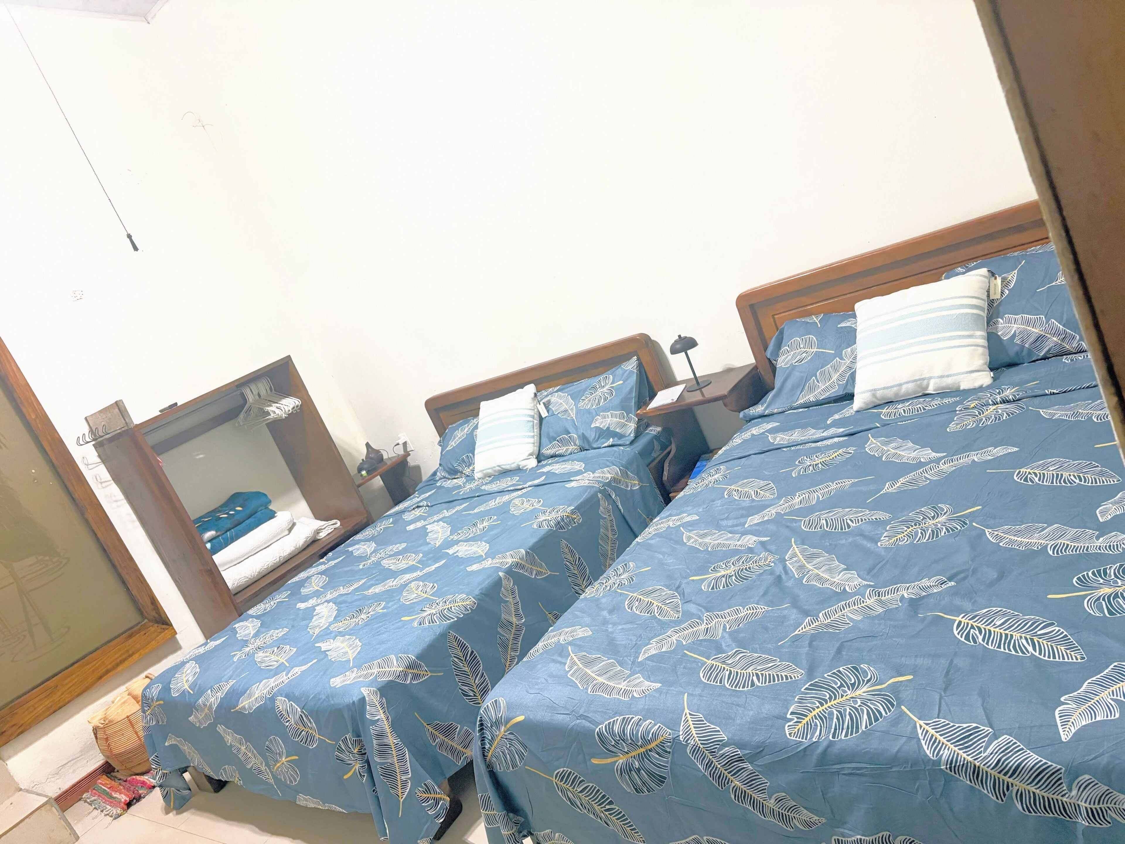 A bedroom with two beds, each with a blue bedspread featuring a leaf pattern. There is a wooden headboard, a bedside table with a lamp, and a mirror on the wall.
