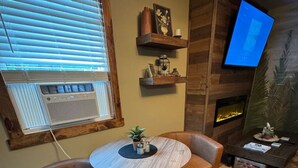 Une pièce avec une table ronde, deux fauteuils en cuir, un plancher en bois, un téléviseur fixé au mur et une cheminée.