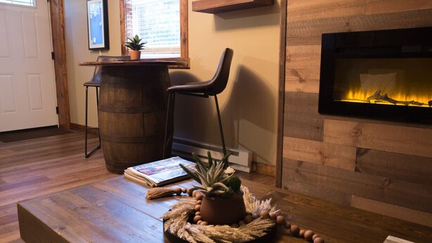 Une pièce chaleureuse avec un plancher en bois, une cheminée, un fauteuil, une table avec une plante et une fenêtre avec des stores.