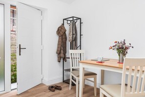 Ein Zimmer mit weißer Tür, einem Kleiderständer mit Jacken, einem Tisch mit Tasse und Blumen sowie einem Schuhpaar auf dem Boden.
