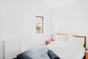 Ein Schlafzimmer mit Bett, Nachttisch, Lampe, Fenster und Heizkörper.