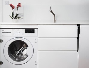 Ein kompakter, weißer Waschraum mit einer Frontlader-Waschmaschine und einem kleinen Blumentopf an der Wand.