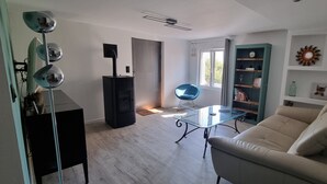 En stue med sofa, sofabord i glas, en blå lænestol, reol og pejs.