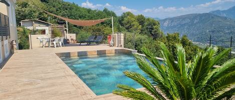 A pool area with a wooden deck, lounge chairs, and a view of mountains.