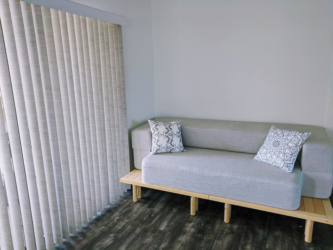 A room with a grey sofa, two patterned pillows, a wooden bench, and a window with vertical blinds.