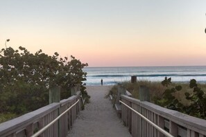 Ein hölzerner Steg führt zu einem Strand, im Hintergrund sind Wellen zu sehen.