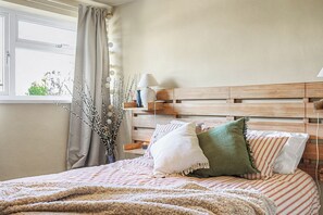 Een slaapkamer met een houten hoofdbord, een raam met gordijnen, een nachtkastje met een lamp, en een bed met gestreept beddengoed en meerdere kussens.
