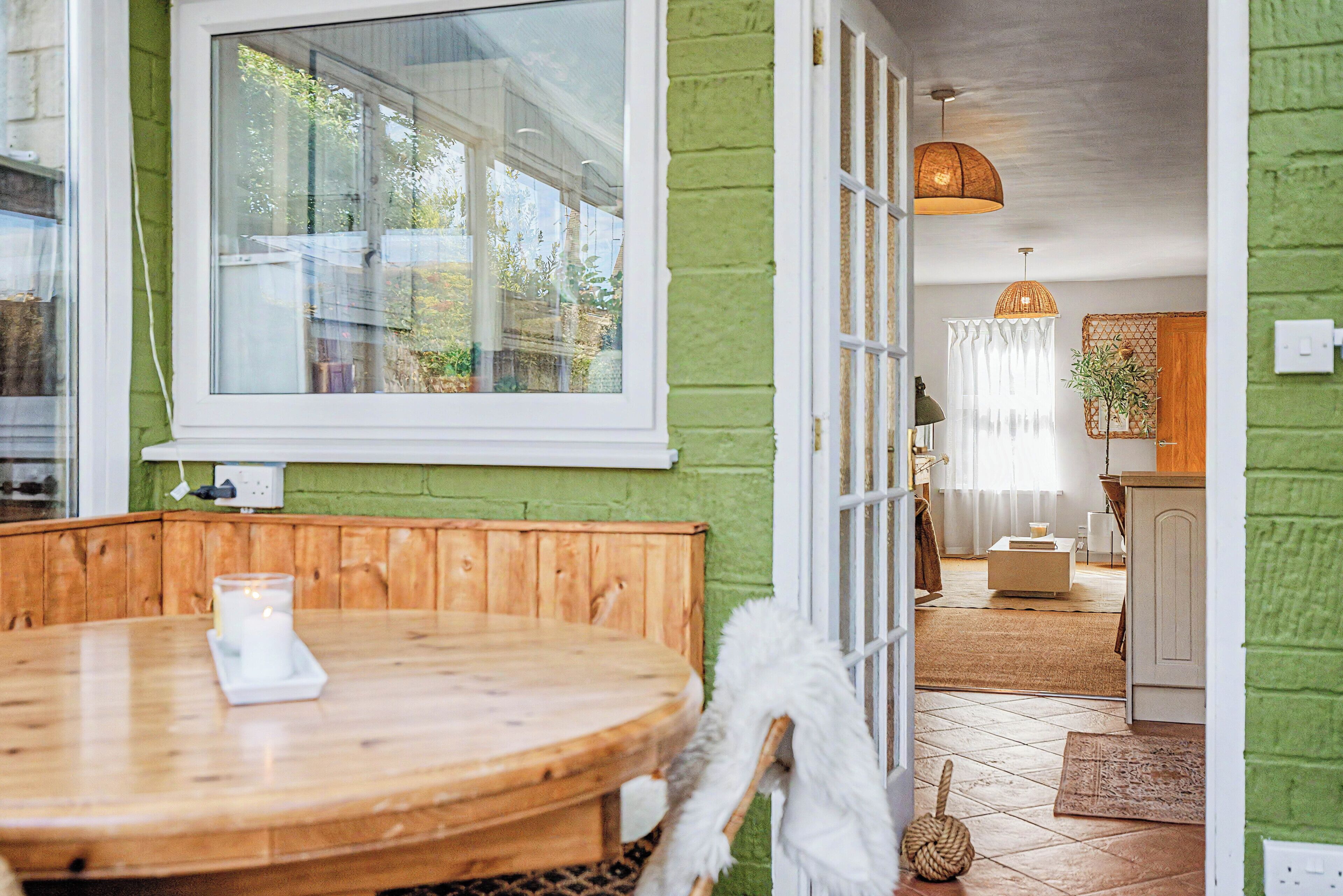 Una habitación con una mesa de madera, una silla blanca, un ventanal grande y una pared verde.