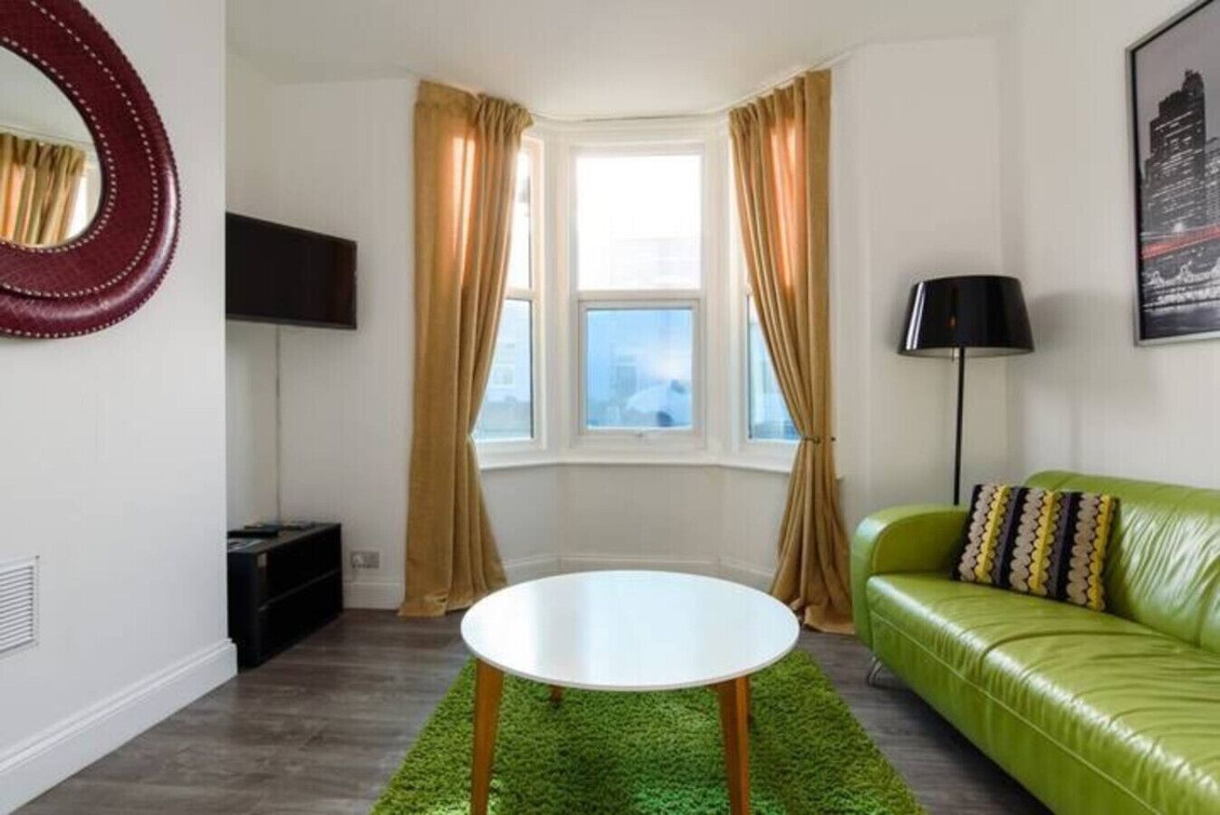 A room with a green sofa, a round white table, a TV on the wall, and a window with curtains.
