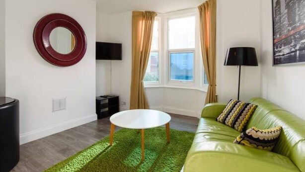 A modern living room with a green sofa, a round coffee table, a red circular mirror, a flat-screen TV, a lamp, and a window with curtains. - Funky Easton flat with parking (Greenbank)