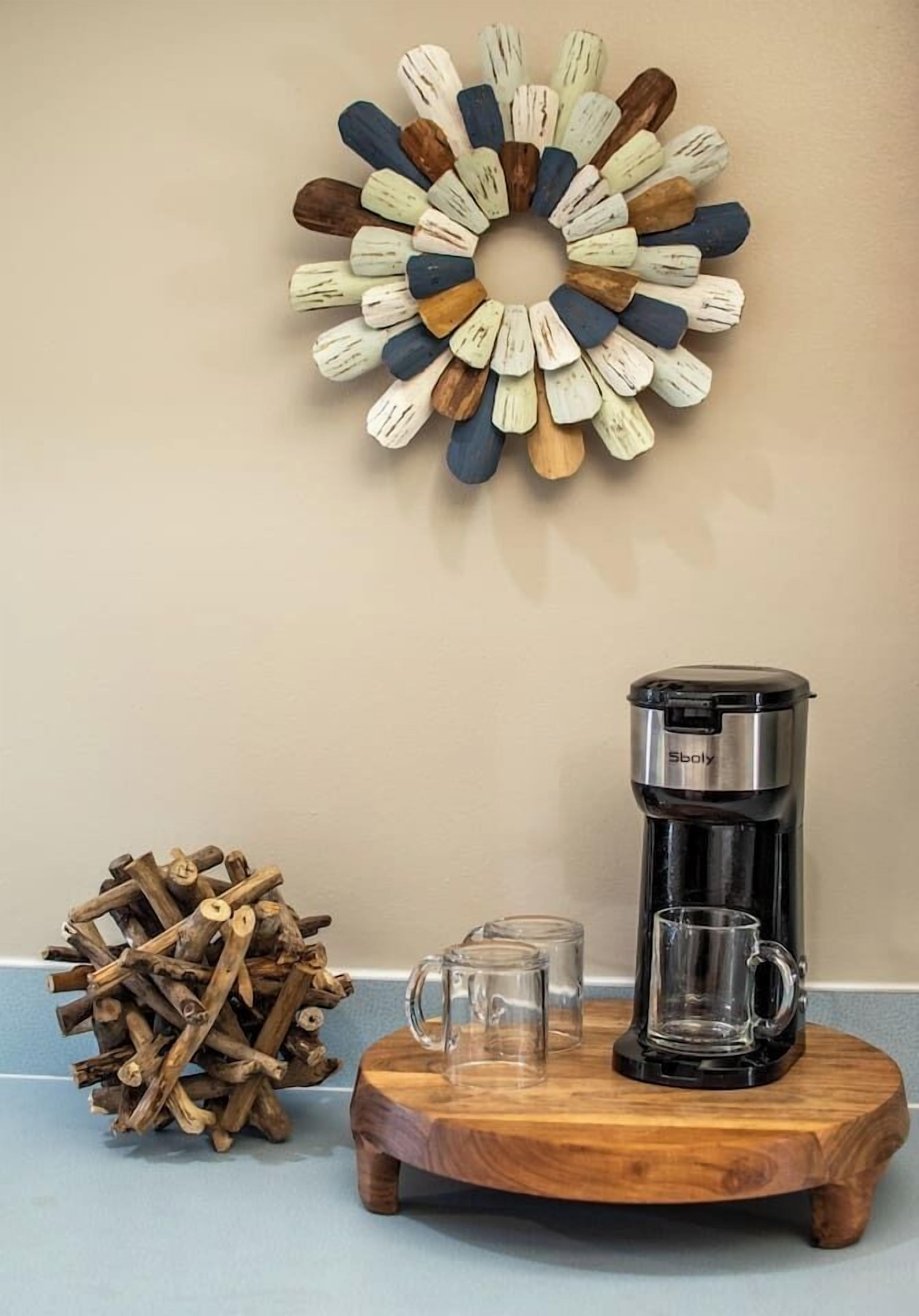 Una cafetera sobre un posavasos de madera, una taza de vidrio y una pila de leña junto a una pared con un arreglo decorativo de madera.