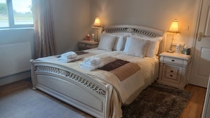 A neatly made bed with a white headboard, two bedside tables with lamps, and a view of the outdoors through a window. - St John s B and B (Lecarrow)