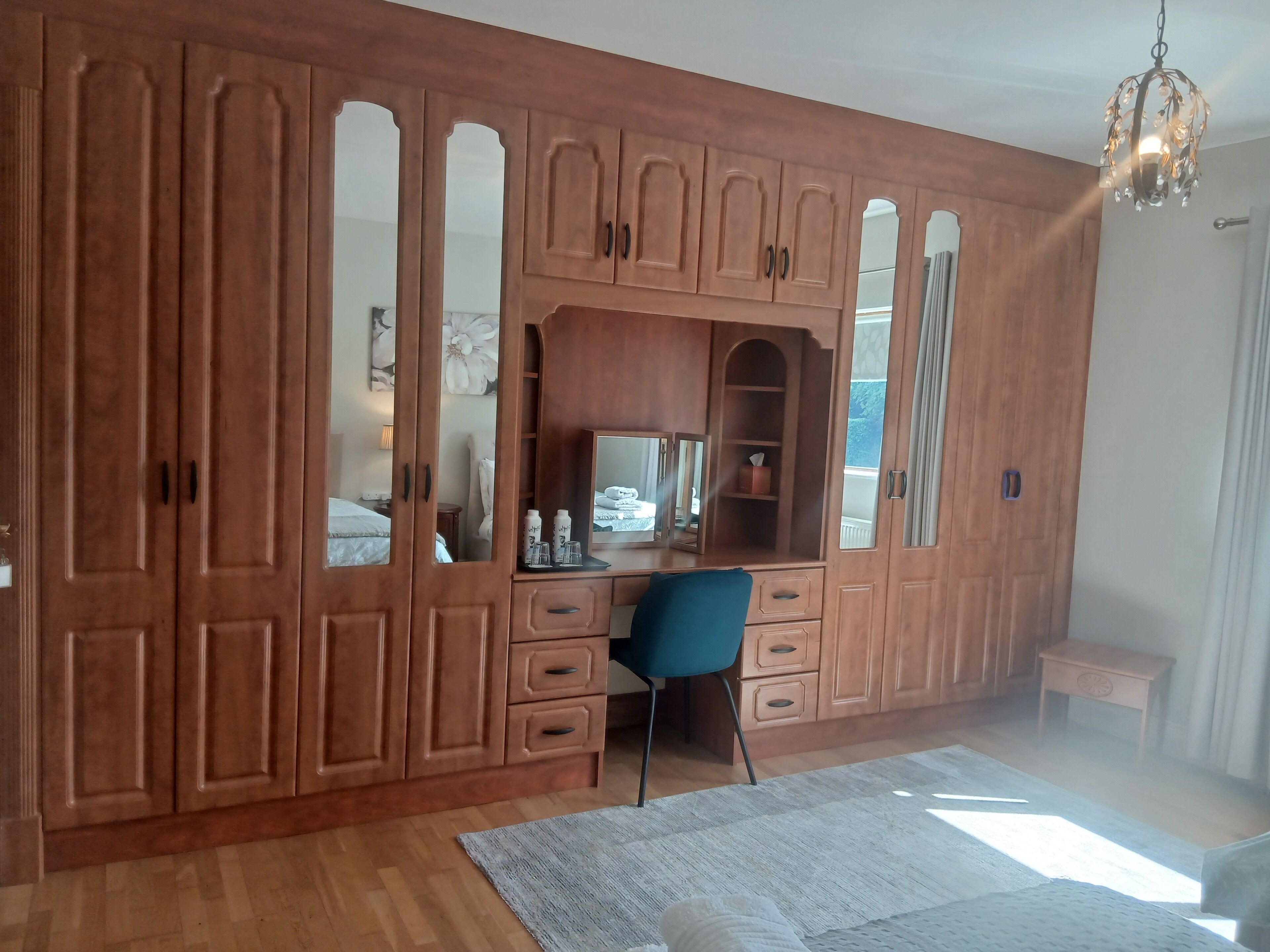A room with a large wooden wardrobe, a desk with a chair, a mirror, and a hanging light fixture.