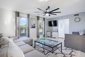 A modern living room with a sofa, a glass coffee table, a ceiling fan, and a TV mounted on the wall.