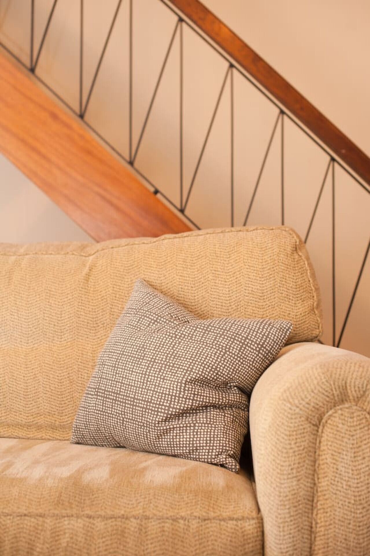 A beige sofa with a patterned pillow against a wall with a wooden staircase.