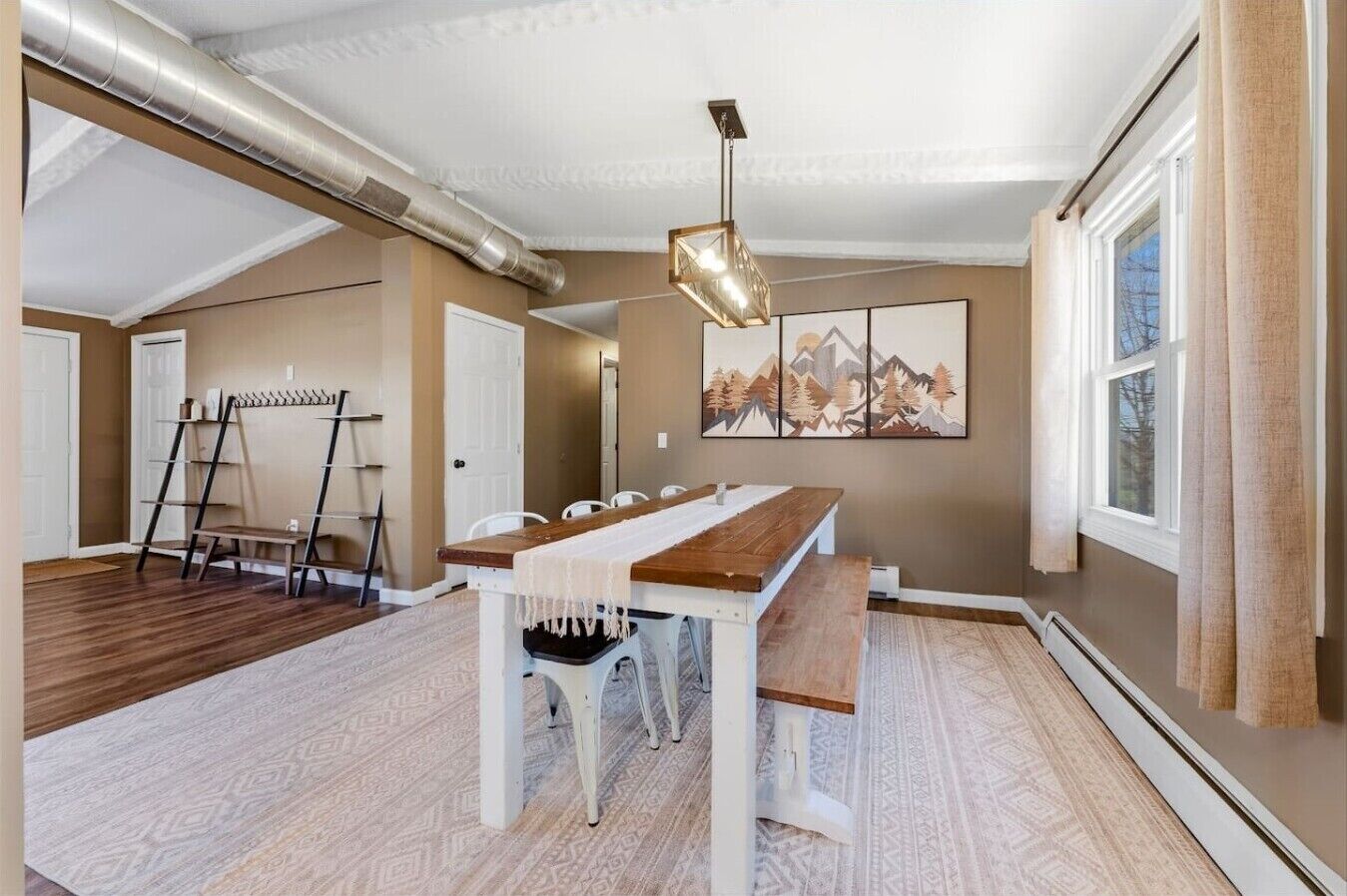Ein geräumiger Raum mit einem langen Holztisch, einer Hängeleuchte und einem Berglandschaftsbild an der Wand.