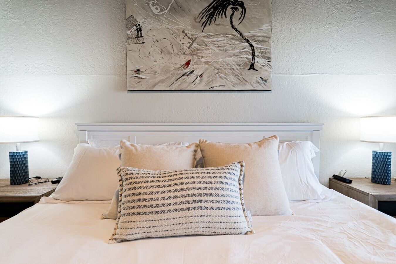 Un lit bien fait, avec du linge de lit blanc et des coussins à motifs, une grande peinture abstraite au-dessus de la tête de lit, et deux lampes de chevet posées sur des tables de nuit en bois.