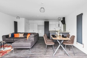 A modern living space with a grey sectional sofa, a dining table with four chairs, and a kitchen area with white cabinets and a grey countertop. - Garden View by MyCityHaven (Redland)