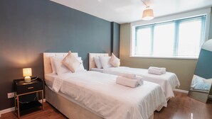 Une chambre avec deux lits, une table de chevet avec une lampe, une fenêtre avec des stores et un miroir.