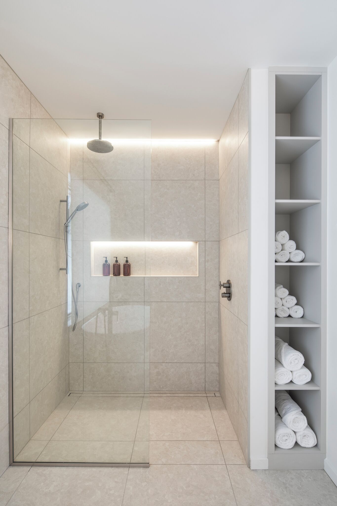 A modern bathroom with a glass-enclosed shower, a built-in shelf with toiletries, and a towel rack with neatly rolled towels.