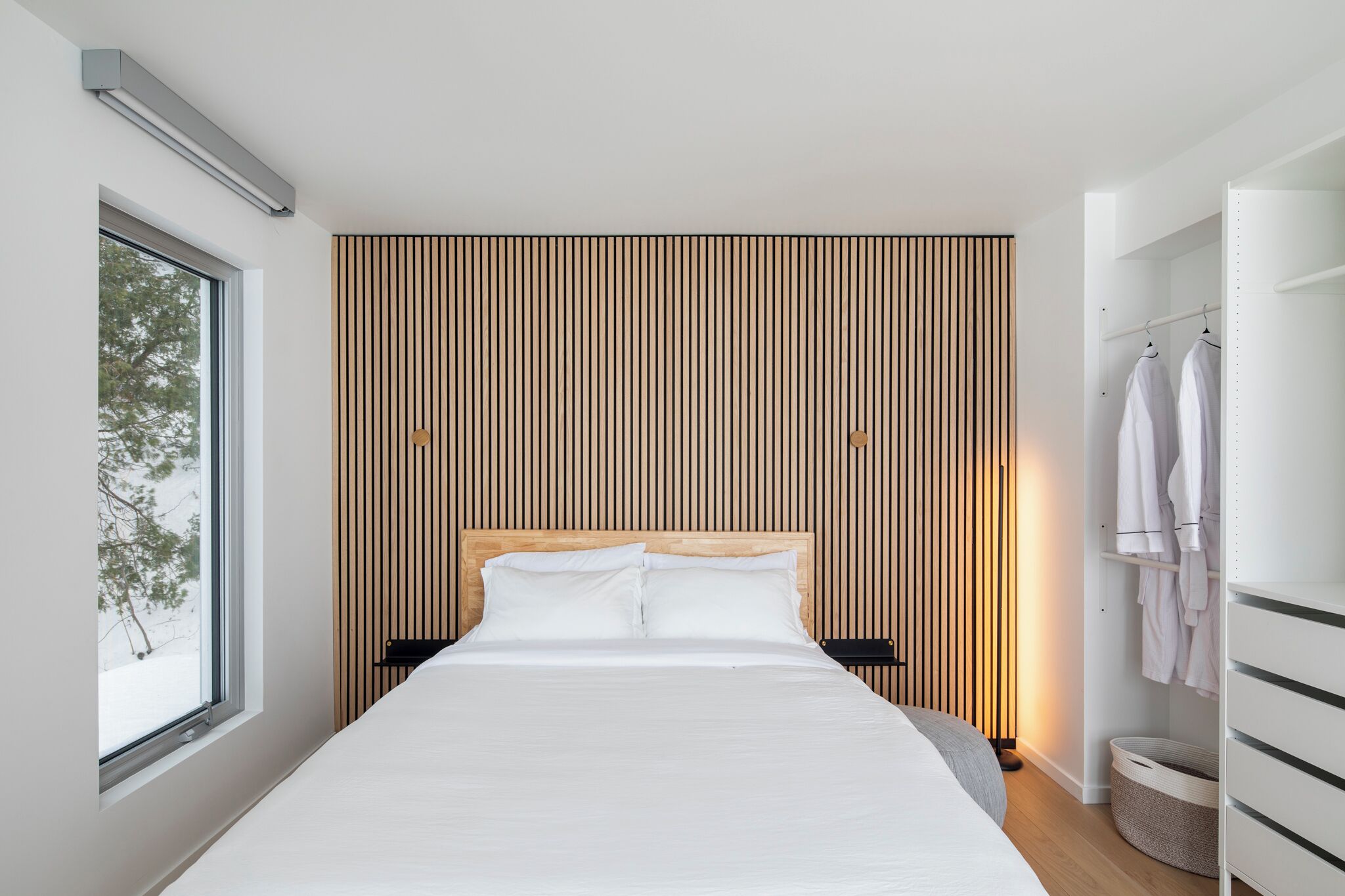 A modern bedroom with a large bed, a wooden slat wall, a window, and a wardrobe.