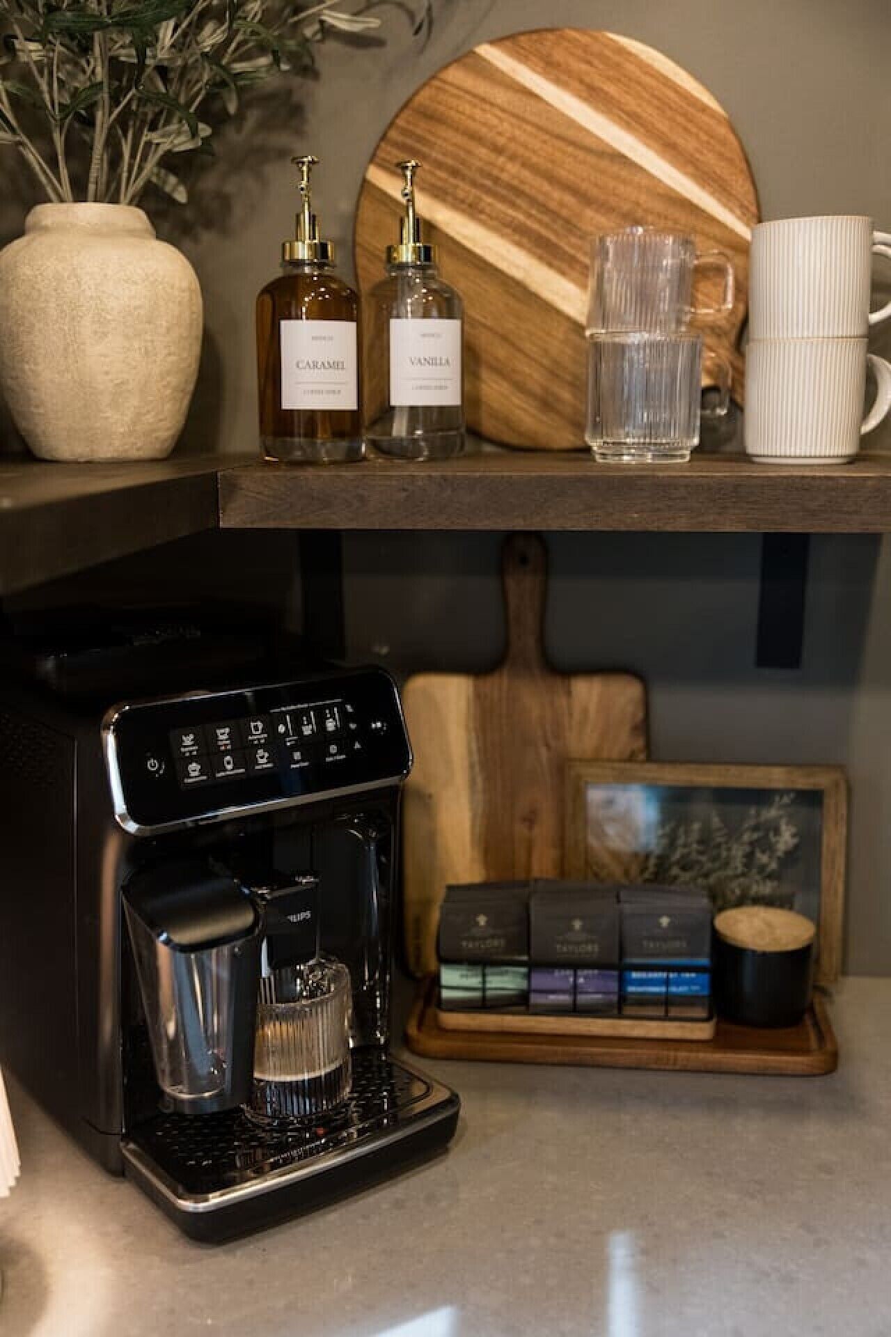 A coffee machine, two bottles of syrup, a glass mug, and a wooden tray with coffee pods.