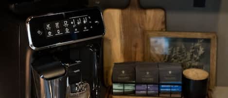 Una cafetera, dos botellas de sirope, una taza de vidrio y una bandeja de madera con cápsulas de café.