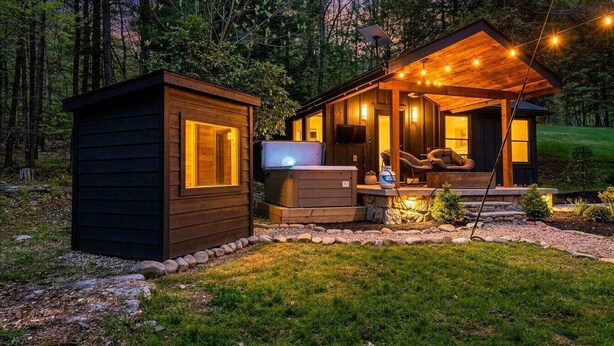 Una cabaña con un cobertizo pequeño adosado, un jacuzzi y una zona de estar cubierta con luces de cuerda.