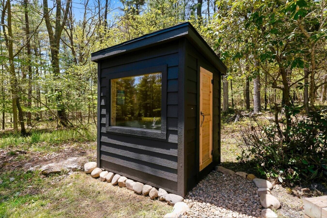 Una petita cabana de fusta de color fosc amb una finestra gran, envoltada d'arbres i un camí de grava.