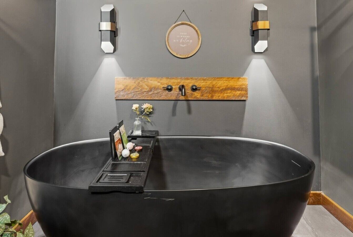 Un baño moderno con una bañera negra grande, un estante de madera con un letrero enmarcado y apliques de pared.