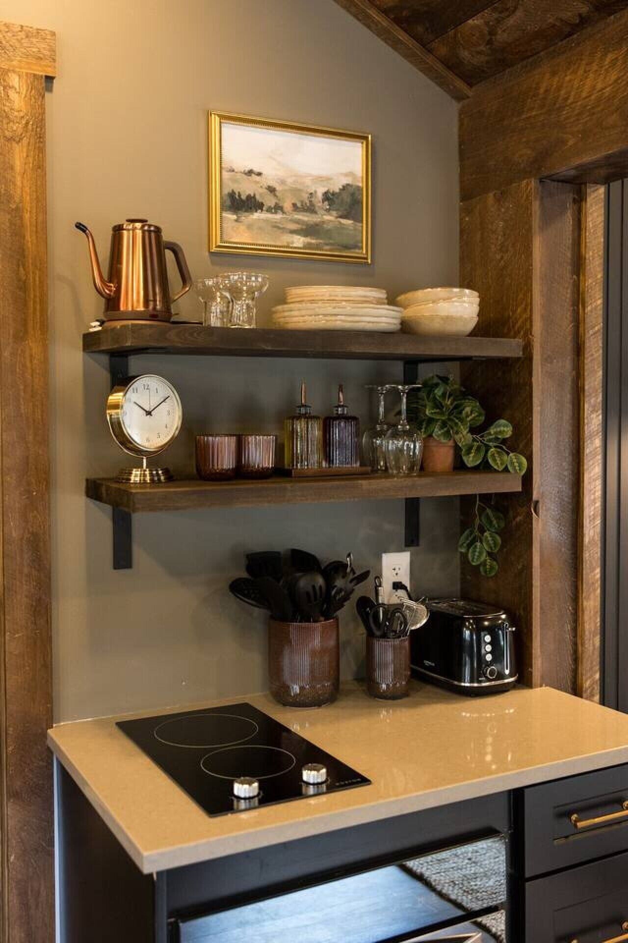 A kitchen with a stove, shelves with kitchenware, a clock, and a framed landscape painting.