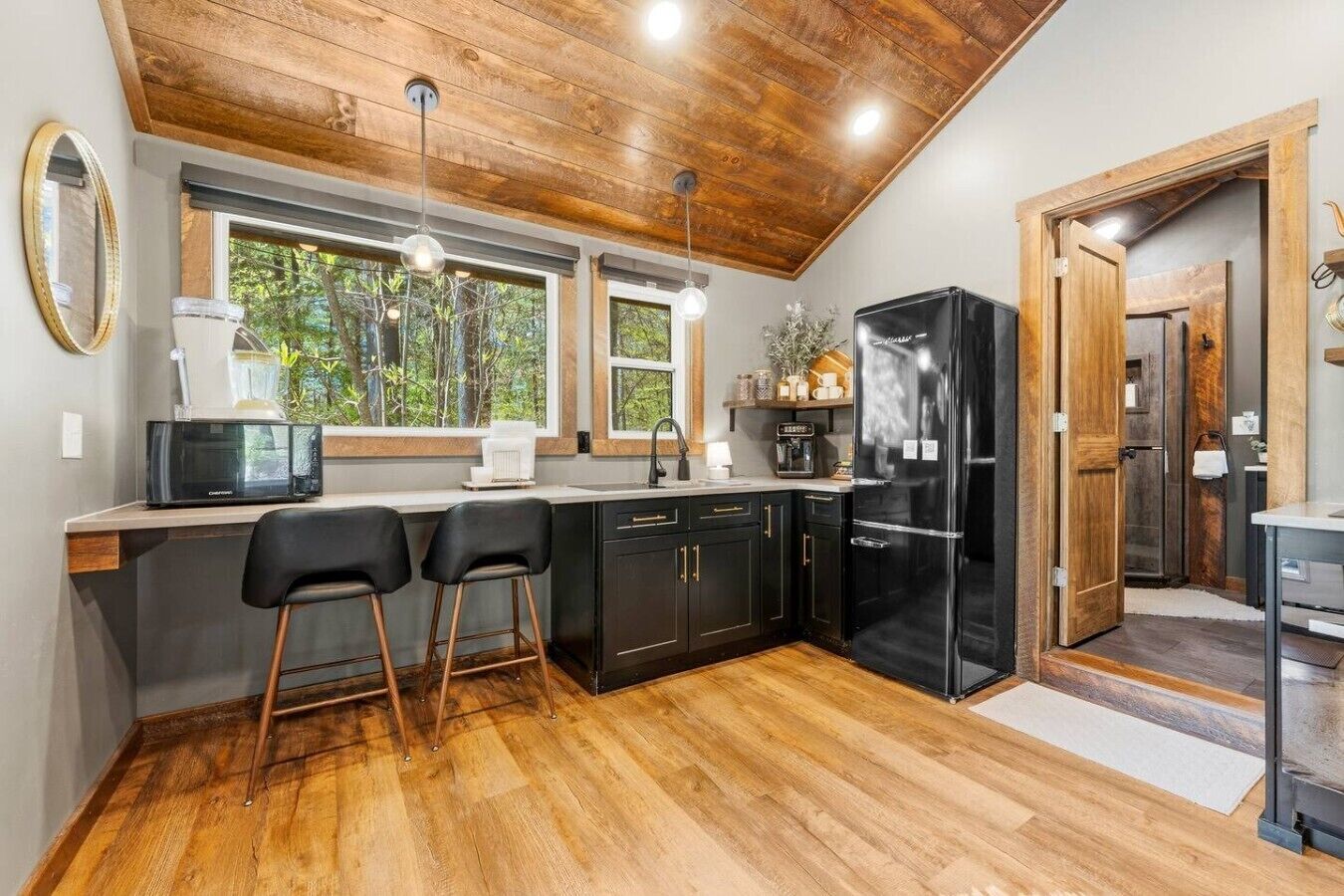 Une cuisine moderne avec un plafond en bois, des meubles de couleur noire, un réfrigérateur noir et une fenêtre donnant sur un paysage verdoyant.